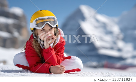 Five-year-old girl in a red-white ski suit and yellow helmet sits thoughtfully on a snow-covered mountain, enjoying the winter landscape 133711927