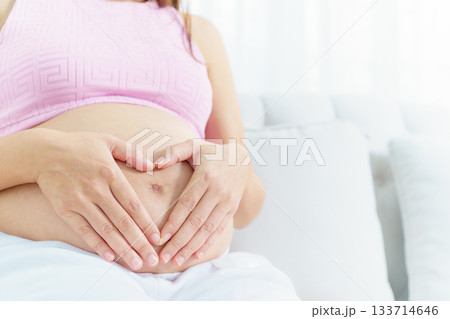 Asian pregnant woman relaxing at home. Asian pregnant woman is sitting and relaxing on a sofa in living room and putting her hands on stomach in the heart shape. 133714646