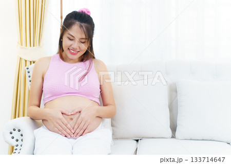 Asian pregnant woman relaxing at home. Asian pregnant woman is sitting and relaxing on a sofa in living room and putting her hands on stomach in the heart shape. 133714647
