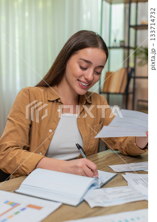 Young woman reviewing receipts nodding satisfied after accurate calculations writing notes at desk Young woman reviewing receipts nodding satisfied after accurate calculations writing notes at desk 133714732