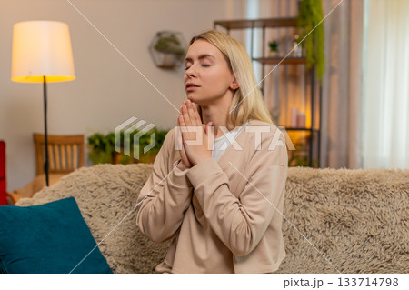 Adult woman praying at home on sofa, folding hands looking upward with sincere emotional expression Adult woman praying at home on sofa, folding hands looking upward with sincere emotional expression 133714798