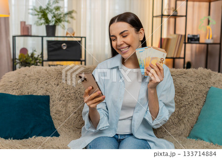 Young woman on sofa holding euro cash and smartphone thoughtfully planning spending choices 133714884