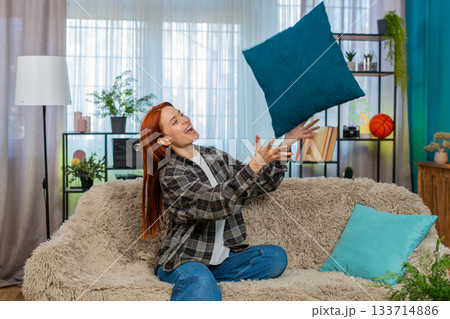 Young woman on sofa playfully tossing pillow in air smiling with joy and cheerful relaxed emotion 133714886