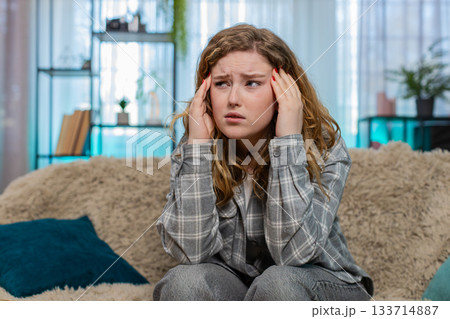Woman sitting on sofa holding head suffering from strong headache or migraine feeling unwell pain 133714887