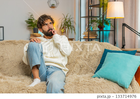 Sad lonely Indian young man sitting on sofa at home room looks pensive thinks over life concerns 133714976