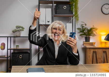 Happy mature elderly woman using smartphone typing browsing celebrating success victory at home 133714991