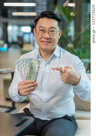 Mature businessman with dollar banknotes showing thumbs up and looking at camera in office lobby Mature businessman with dollar banknotes showing thumbs up and looking at camera in office lobby 133715020