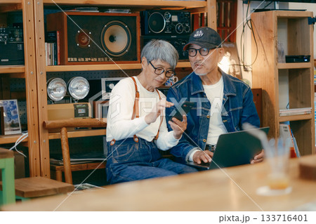 シニア夫婦がカフェでパソコンとスマホを使って調べものをするシーン 133716401