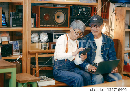 シニア夫婦がカフェでパソコンとスマホを使って調べものをするシーン 133716403