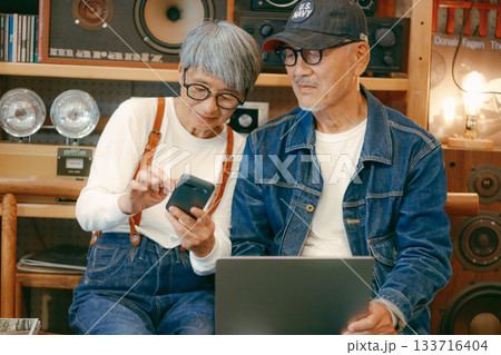 シニア夫婦がカフェでパソコンとスマホを使って調べものをするシーン 133716404