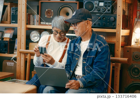 シニア夫婦がカフェでパソコンとスマホを使って調べものをするシーン 133716406