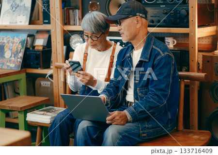 シニア夫婦がカフェでパソコンとスマホを使って調べものをするシーン 133716409