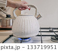 A person is holding a white tea kettle over a gas stove 133719951