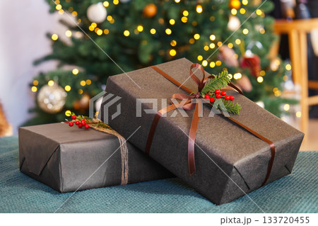Two Christmas gift boxes decorated with ribbon and red holly berry in front of glowing festive tree Two Christmas gift boxes decorated with ribbon and red holly berry in front of glowing festive tree 133720455