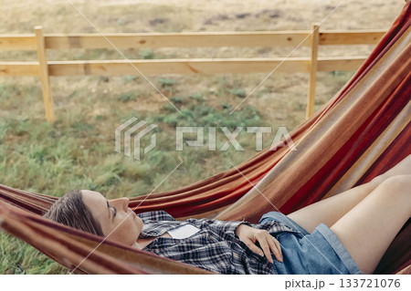 young Caucasian woman with long brown hair relaxes in a colorful hammock under a tree in a rural setting. The scene conveys a sense of tranquility and escape young Caucasian woman with long brown hair relaxes in a colorful hammock under a tree in a rural setting. The scene conveys a sense of tranquility and escape 133721076