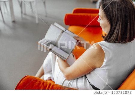 young Hispanic woman with short brown hair sits on an orange couch in a library, reading a book. Bookshelves filled with books are visible in the background. 133721078