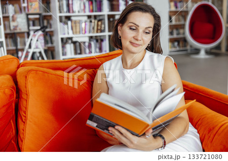 Hispanic woman with short brown hair sits on an orange couch in a library, reading a book. Bookshelves filled with books are visible in the background. 133721080