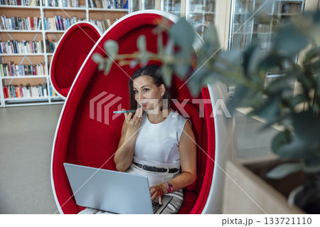 young Caucasian woman with brown hair sits in a red egg chair, using a laptop and talking on a phone in a modern coworking space. young Caucasian woman with brown hair sits in a red egg chair, using a laptop and talking on a phone in a modern coworking space. 133721110