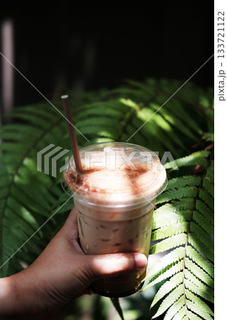 Close up hand holding a plastic glass of ice latte coffee with chocolate powder on topping in natural sunlight and tropical fern forest garden 133721122