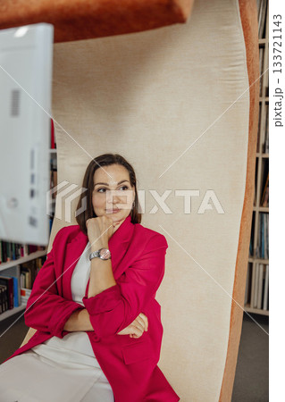 A young Caucasian woman with brown hair sits thoughtfully in a modern coworking space. She wears a red blazer and has a relaxed posture, surrounded by bookshelves. A young Caucasian woman with brown hair sits thoughtfully in a modern coworking space. She wears a red blazer and has a relaxed posture, surrounded by bookshelves. 133721143