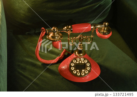 Close-up of a red rotary phone with an elegant brass dialer over velvet surface. phone of vintage design. Red rotary phone with brass dials, its cord placed artfully on soft green velvet. 133721495
