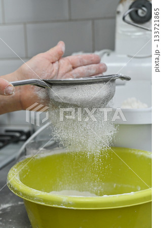 Hand using sieve to sift flour into yellow bowl. Household baking process symbolizing ingredient preparation, dough making. Closeup of flour passing through mesh sieve. vertical Hand using sieve to sift flour into yellow bowl. Household baking process symbolizing ingredient preparation, dough making. Closeup of flour passing through mesh sieve. vertical 133721865