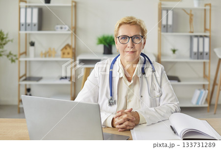 Mature female doctor smiling at desk, medical consultant, female medical nurse or practitioner Mature female doctor smiling at desk, medical consultant, female medical nurse or practitioner 133731289