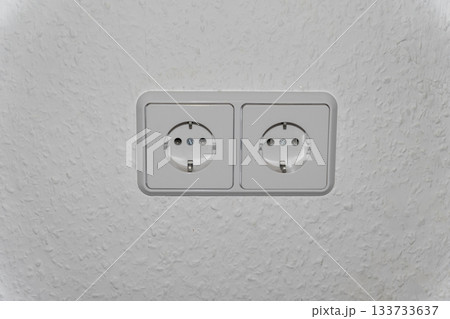 A double electrical outlet (Schuko type) in white or light gray is centered on a plain light-colored wall. The image focuses on themes of electricity, renovation, construction, home electrical systems 133733637