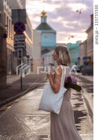 A woman stands on a wet city street at sunset with a white canvas tote and pink hydrangea, framed by soft pastel tones and historic buildings 133735580