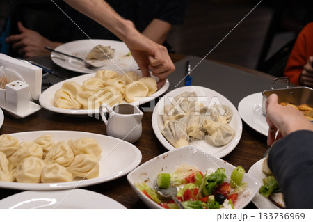 In a cozy setting, hands reach for steaming plates of dumplings and fresh salad as family members gather around. The atmosphere is filled with warmth, taste, and connection. 133736969