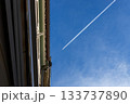An airplane leaving a white contrail across a bright blue sky, viewed from below between two buildings, creating a dynamic urban travel scene. 133737890