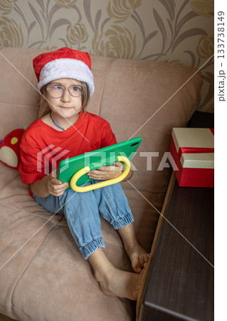 A girl in a Santa Claus hat opens a gift. It's Christmas time. A girl in a Santa Claus hat opens a gift. It's Christmas time. 133738149
