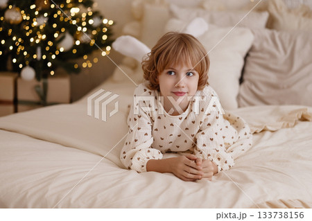 Caucasian child lying on bed in cozy bedroom with Christmas tree, look at camera Caucasian child lying on bed in cozy bedroom with Christmas tree, look at camera 133738156