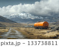 Orange tanker trailer resting in a wide valley with snowy mountains under dramatic clouds 133738811