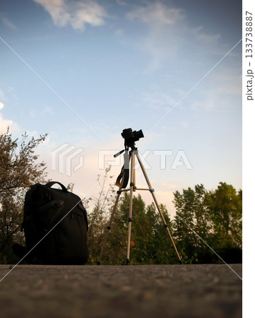 silhouette of a backpack and camera on a tripod against the sky and green trees at dusk silhouette of a backpack and camera on a tripod against the sky and green trees at dusk 133738887