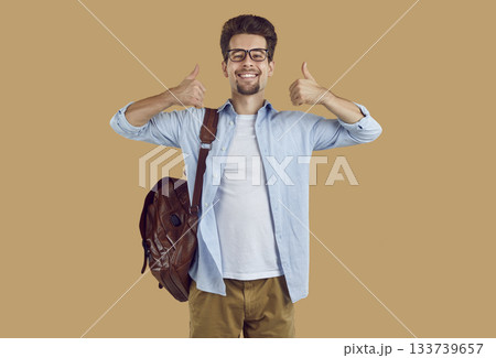 Happy university student guy with leather backpack showing thumbs up on beige background. 133739657