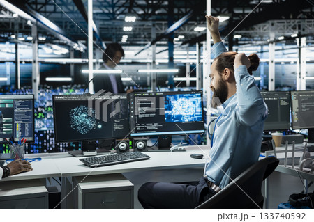Data center system administrator stretching at PC desk to relieve back fatigue while using AI assistant to perform maintenance. Server room employee relaxing sore muscles after long hours at computer 133740592