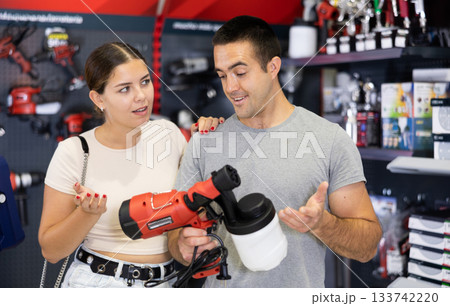 In store, spouses buyer doubts choice and closely examines electric paint-spraying machine spray gun In store, spouses buyer doubts choice and closely examines electric paint-spraying machine spray gun 133742220