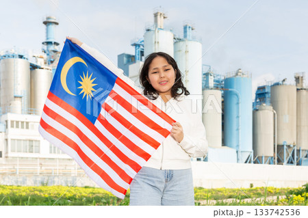 Happy young woman with flag of Malaysia against background of factory Happy young woman with flag of Malaysia against background of factory 133742536