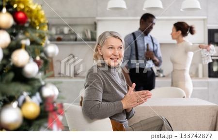 Smiling old woman sitting at the kitchen table with her back to couple quarreling in the kitchen with Xmas-tree Smiling old woman sitting at the kitchen table with her back to couple quarreling in the kitchen with Xmas-tree 133742877