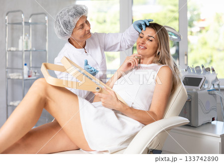 Young woman looking in mirror while experienced cosmetologist explaining facial procedure 133742890