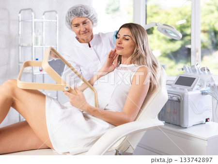 Young woman looking in mirror while mature cosmetologist showing results of facial procedure 133742897