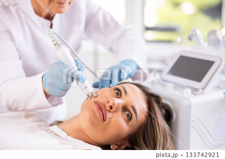 Young female client receiving hardware ultrasonic to revitalize and tighten facial skin at clinic Young female client receiving hardware ultrasonic to revitalize and tighten facial skin at clinic 133742921