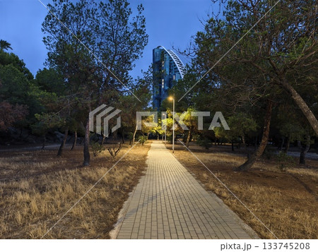 quiet dusk pathway beside tall glass building, serene evening walk through shimmering lamppostlit 133745208