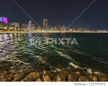 night sea scene, rocky shore with city lights, coastal night scene with urban illumination 133745255