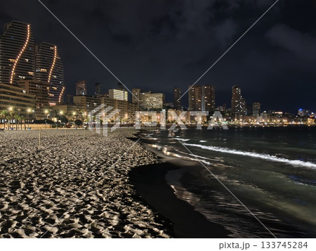 foamy shoreline at night, waves softly lapping illuminated shoreline with city backdrop at night 133745284