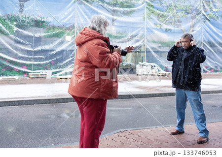 Female photographer in red jacket filming man in headphones listening to music in snowy rainy weather outside modern business center. High quality photo Female photographer in red jacket filming man in headphones listening to music in snowy rainy weather outside modern business center. High quality photo 133746053