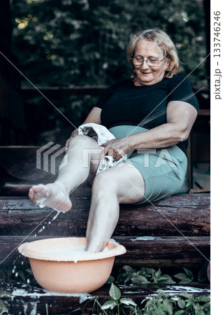 Elderly woman in glasses washing her feet in basin of foam in backyard on hot summer day. High quality photo 133746246