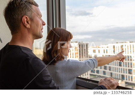 Man hugging his wife. Young couple moving in new apartmen. People looks out window at new houses. Concept new homes, new beginning. High quality photo Man hugging his wife. Young couple moving in new apartmen. People looks out window at new houses. Concept new homes, new beginning. High quality photo 133746258