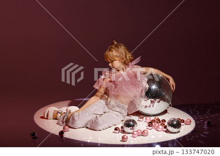 Studio portrait of young girl in festive outfit with disco balls on burgundy background Studio portrait of young girl in festive outfit with disco balls on burgundy background 133747020